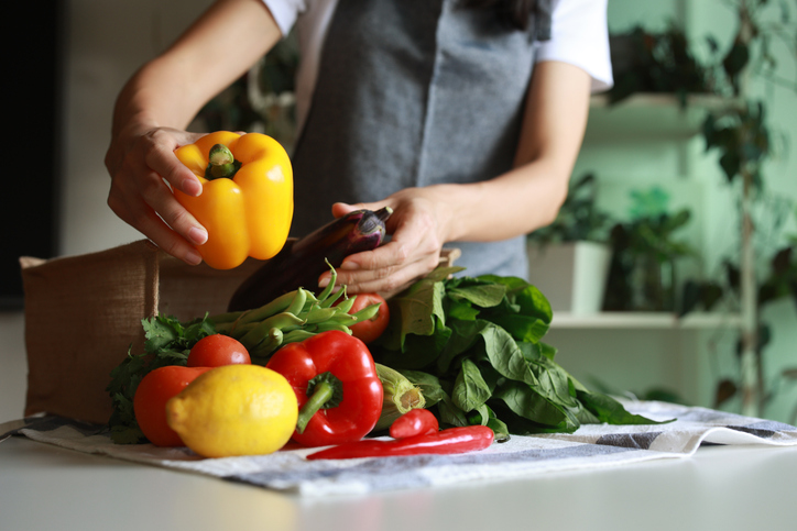 A young Asian woman returns home from grocery shopping, unpacking fresh and healthy organic fruits and vegetables from a reusable bag onto the table. This reflects responsible shopping, zero waste, and a sustainable lifestyle concept.