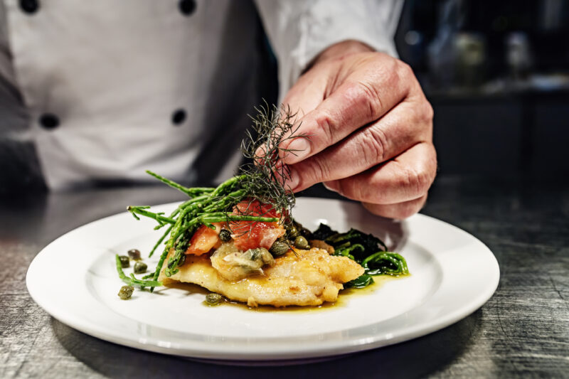 Een chef-kok presenteert een visgerecht met gebakken visfilet, groenten, kappertjes en kruiden op een wit bord.