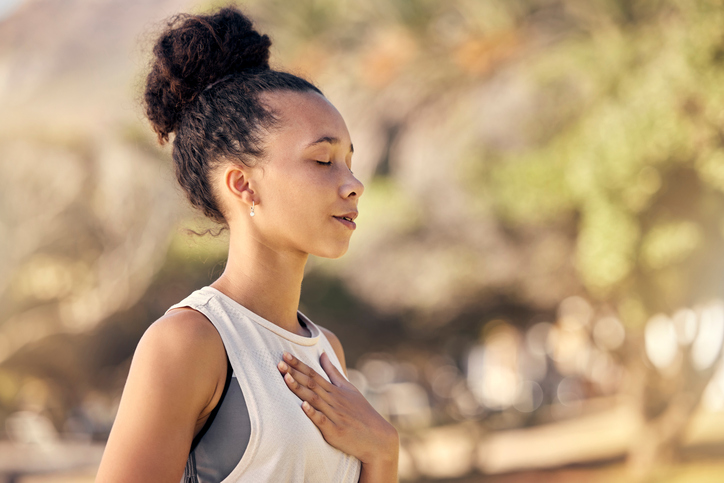 Een vrouw met een serene uitdrukking staat buiten en legt haar hand op haar borst terwijl ze haar ogen sluit en diep ademhaalt.